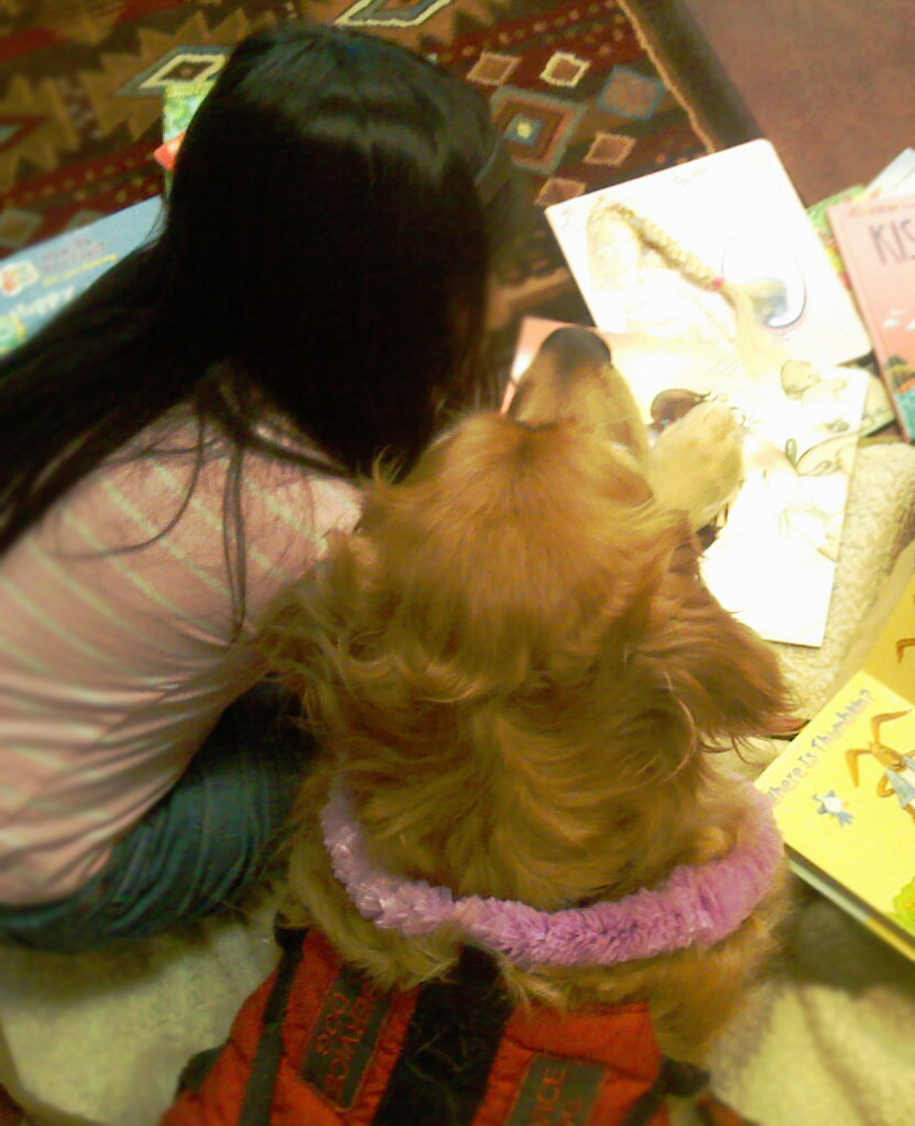 A young girl with long black hair, wearing a pink and white striped shirt, "reads" books with a golden retriever pup.