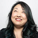 Headshot of a Korean woman with long black hair wearing a black top, smiling and gazing upward