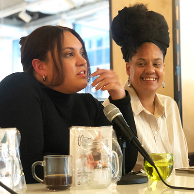 A woman at a table talking into a microphone while another woman looks on.