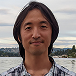 Headshot of a smiling Asian man with black hair, wearing a plaid button-down shirt, in front of a body of water.
