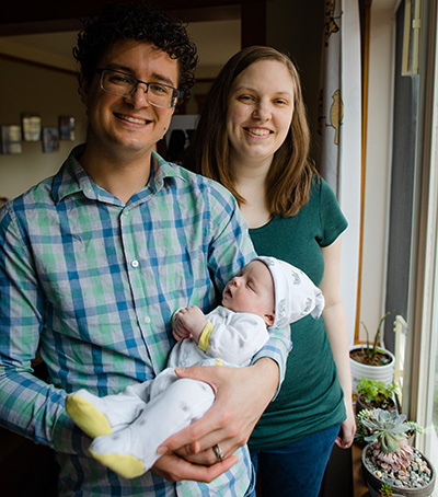 A man in a blue plaid shirts stands besides a woman in a green t-shirt cradling a baby in his arms.