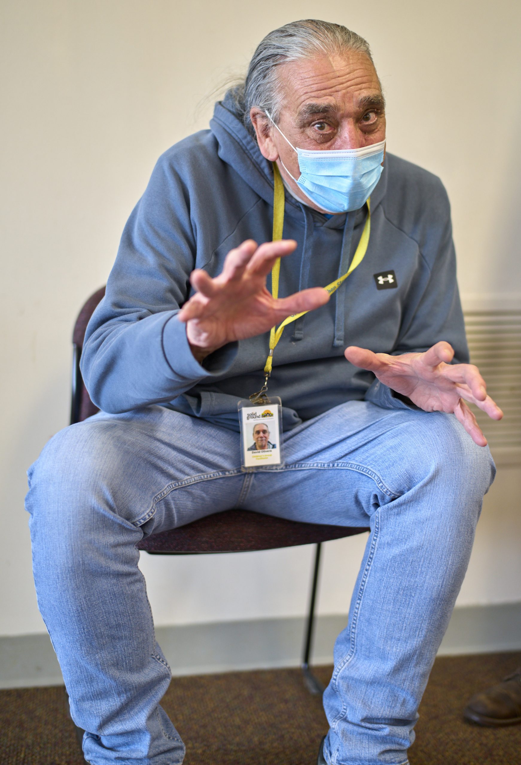 A man with long gray hair and jeans leans forward in a chair and gestures with his hands.