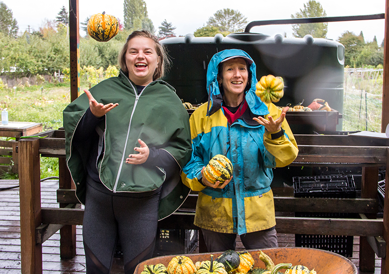 Two women in rain jackets laugh as they toss colorful winter squash from a wheel barrow into the air.