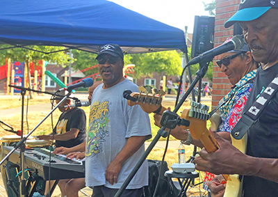 Several men stand before microphones as they perform an outdoor concert. A playground can be seen behind them.