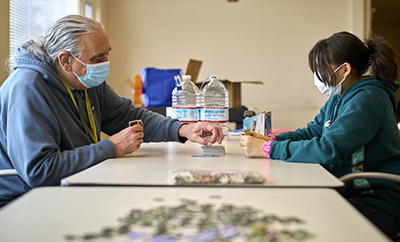 A man with long gray hair and a blood hoodie plays cards with a young woman with dark hair and a turquoise hoodie.