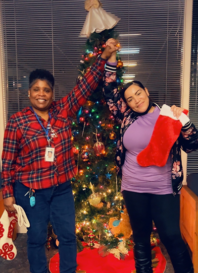 Two women clasp hands in front of a brightly decorated Christmas Tree.