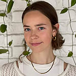 Headshot of a young white woman with dark brown hair, wearing a white top, in front of a white brick wall with green vines hanging down.