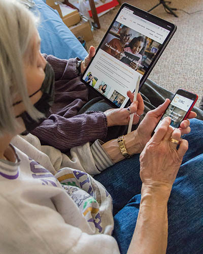 Side shot of a woman in a purple shirt holds a tablet and stylus next to an elderly woman using a cellphone.