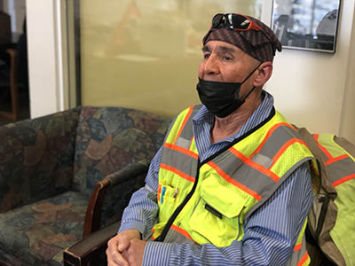 A man with a reflective vest over a blue collared shirt sits in a chair in an office.
