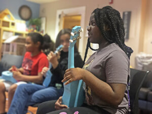 Three children sit on chairs holding blue ukuleles.