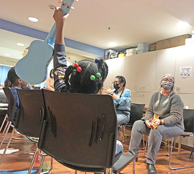 A young girl holds a ukulele above her head, showing her fret fingering to two teachers who can be seen in the background.