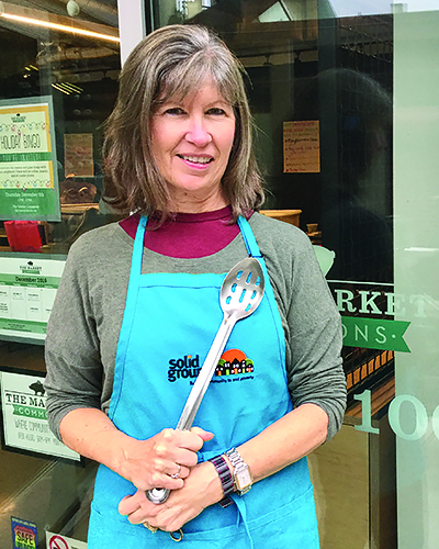 A smiling woman wearing a gray and maroon long-sleeved top and a blue apron holds a slotted spoon.