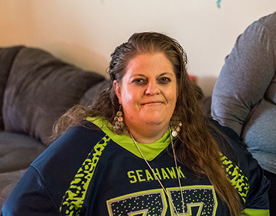 A woman in a Seahawks jersey sits on the arm of a gray couch.