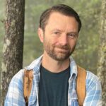 A smiling man in a blue plaid shirt with a forest scene behind him.