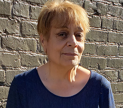 A portrait of a woman with reddish hair and a blue blouse in front of a gray brick wall.