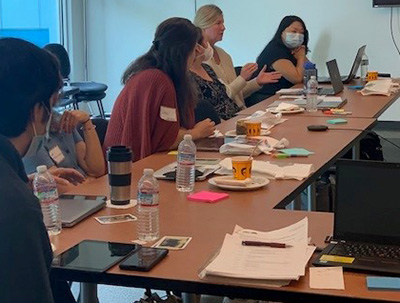 A diverse group of six people sit around an L-shaped table covered with note bads, sticky notes, bottles of water, and laptops.