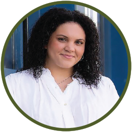 Circular photo with a forest-green frame of a young Black woman with shoulder-length hair, wearing a white blouse, standing in front of a blue building.