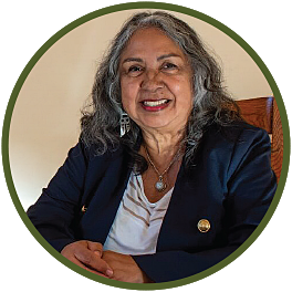 Circular photo with a forest-green frame of a Latina woman with shoulder-length, salt & pepper hair sitting in a wooden-backed chair, wearing a white blouse and dark jacket.