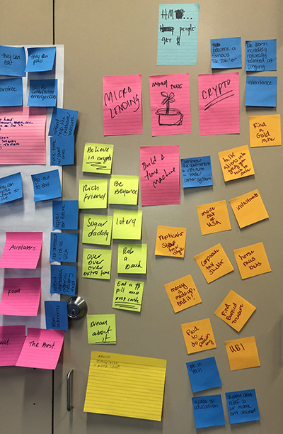 A pair of doors covered in yellow, pink, orange and blue Post It notes with scribbled words and phrases, including "microlending," "crypto," "build a time machine," "sugar daddy," "UBI," "rich friends," and "be Beyonce."