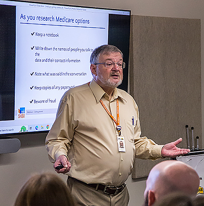 A man with grey hair and beard wearing glasses, an orange lanyard, and khaki pants and shirt, speaks to a small audience. Behind him is a PowerPoint slide titled 'As you research Medicare options.'