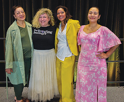 Four women standing and posed shoulder to shoulder, wearing colorful outfits.