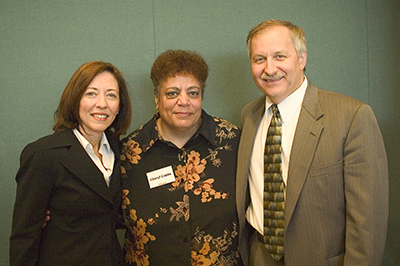 Three people in fancy dress pose together in front of a solid green background.