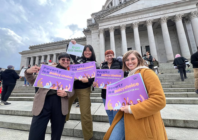 Four people standing on steps infront of a state legislative building holding signs that read "Poverty is a policy choice: Pass Progrressive Revenue For All."