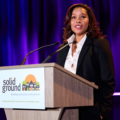 A woman in a dark suit jacket stands at a podium with a sign that reads "Solid Ground."