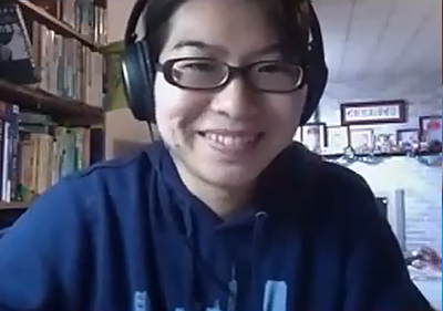 Asian woman with glasses and a blue shirt, wearing headphones, smiles during a Zoom video