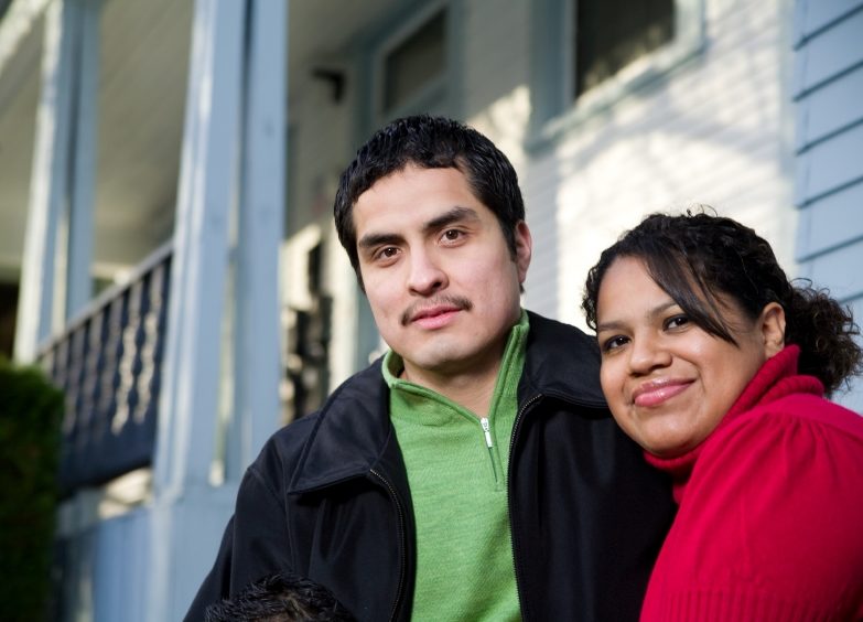Latino man with mustache in a green shirt and jacket and Latina woman in red top smile in front of an apartment building