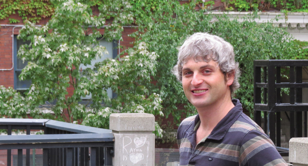 White man with grey hair wearing a striped polo shirt, standing outside in front of a brick building and a green leafy tree