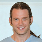 Headshot of a man with dark brown hair in a ponytail, sideburn, and a grey T-shirt