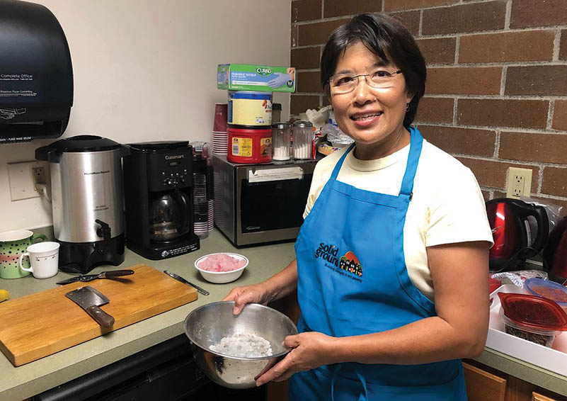 A cooking class volunteer in a blue apron holds a bowl of flour