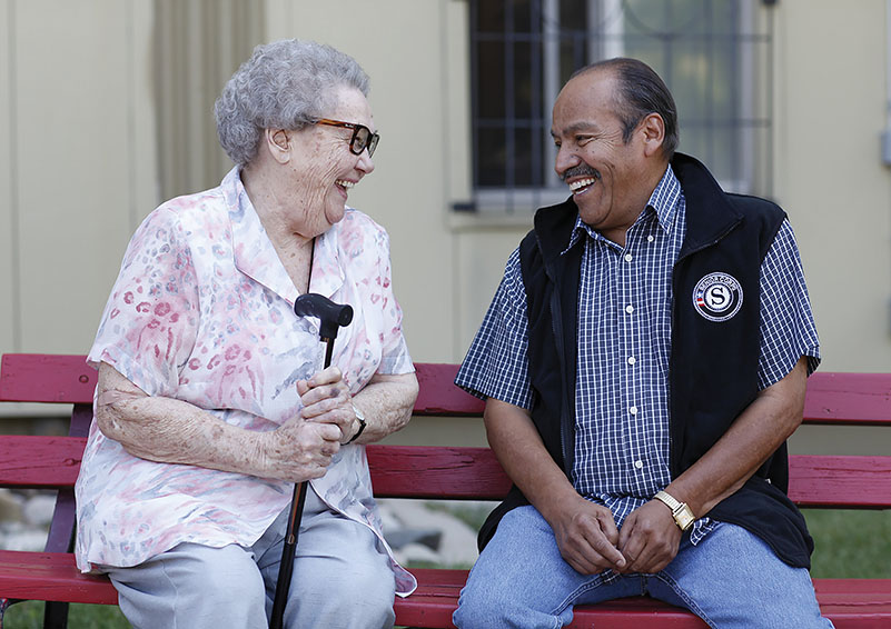Elderly lady with cane sitting next to Senior Volunteer on bench.