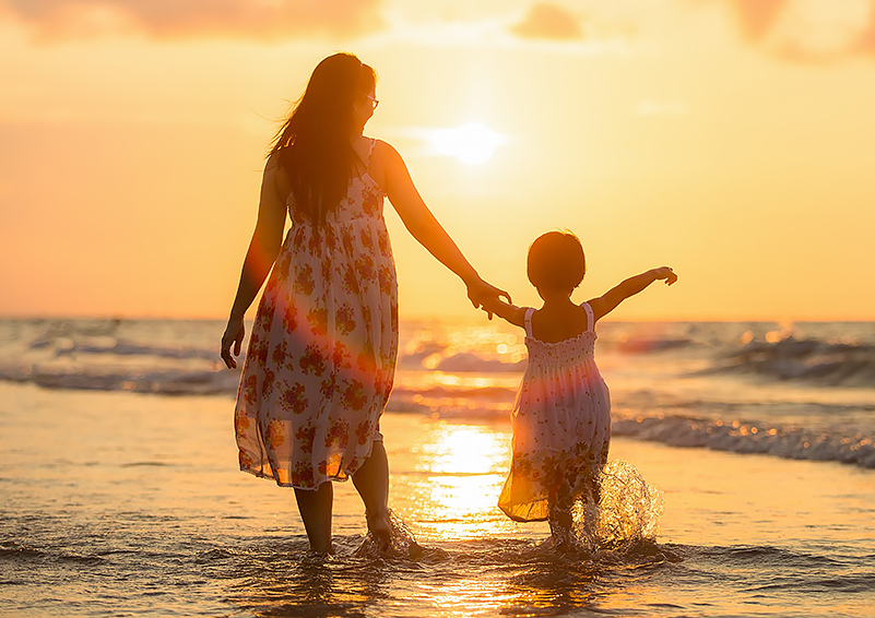 A mother & daughter hold hands and wade into the incoming tide on a beach as the sun begins to go down