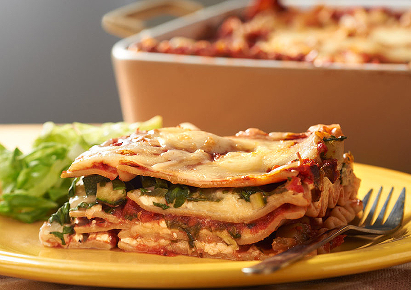 a slice of veggie lasagna and some salad on a yellow plate, in front of a baking pan of lasagna