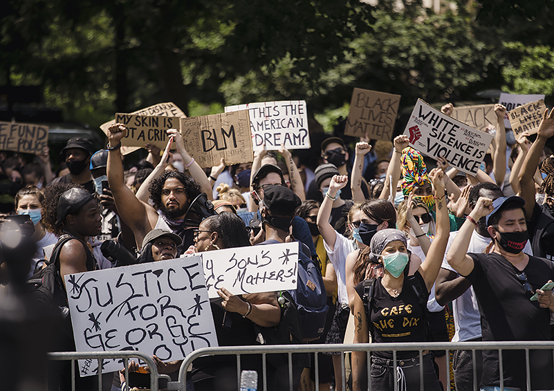 multi-racial crowd of protesters behind a metal barrier fence holding signs: "Justice for George Floyd," "Defund the Police," "BLM," "White Silence = Violence," etc.