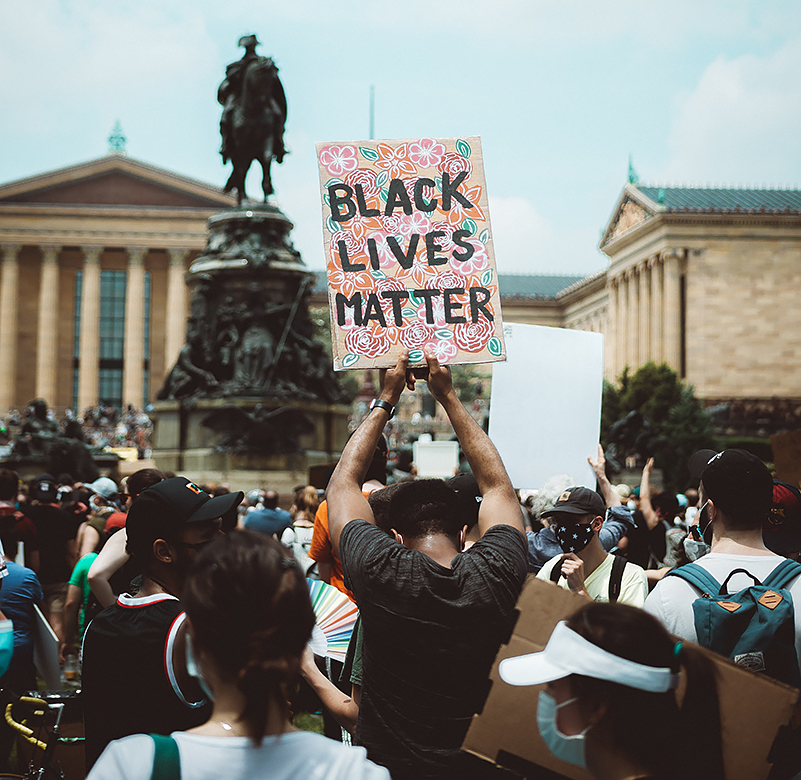 Black man bows his head while holding up a BLACK LIVES MATTER sign (black lettering on an ornate floral background) amidst a crowd, standing in front of a Confederate statue