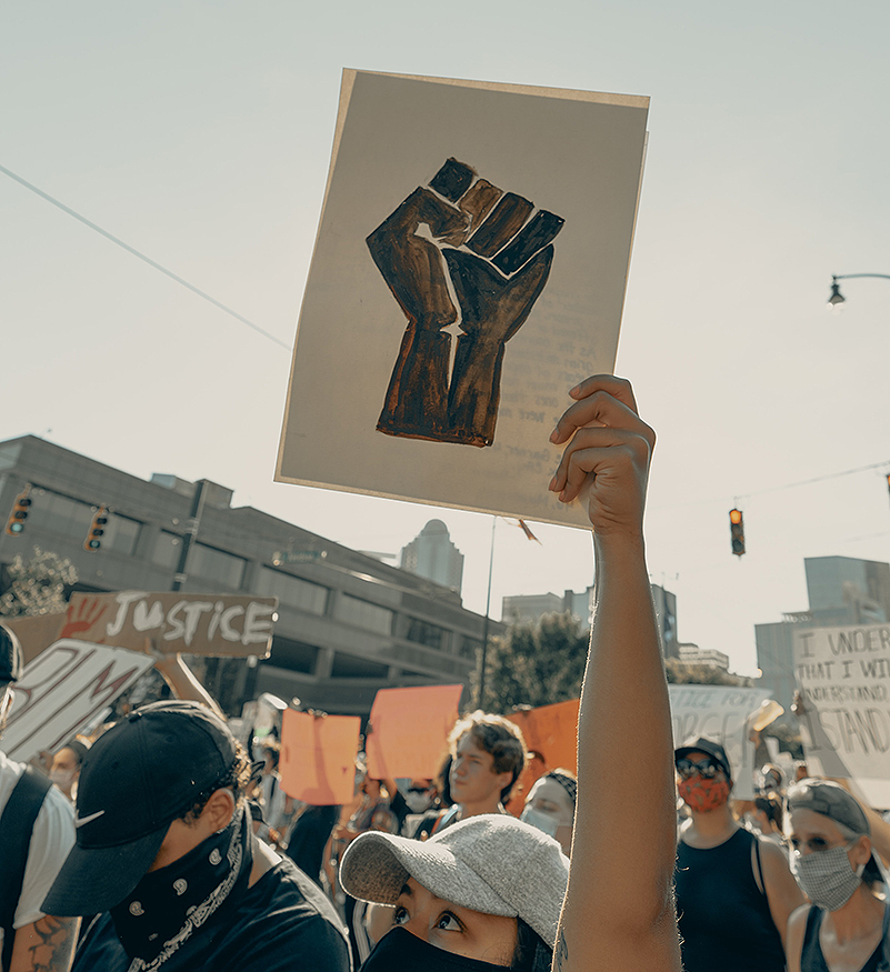 Woman in a white cap & black mask holds up a sign of a Black power fist, no text, amidst a crowd of protesters