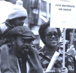 Black and white photo of man in cap with his son on his back, and his wife holding an auto machinists protest sign