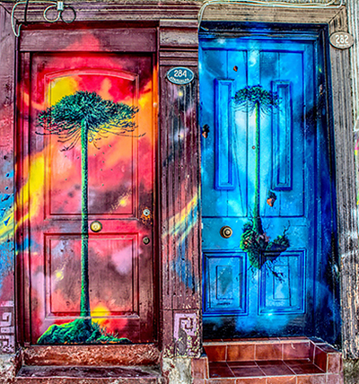 Two brightly painted doors side by side, one pink, red, and yellow with a green tree on it, and one blue