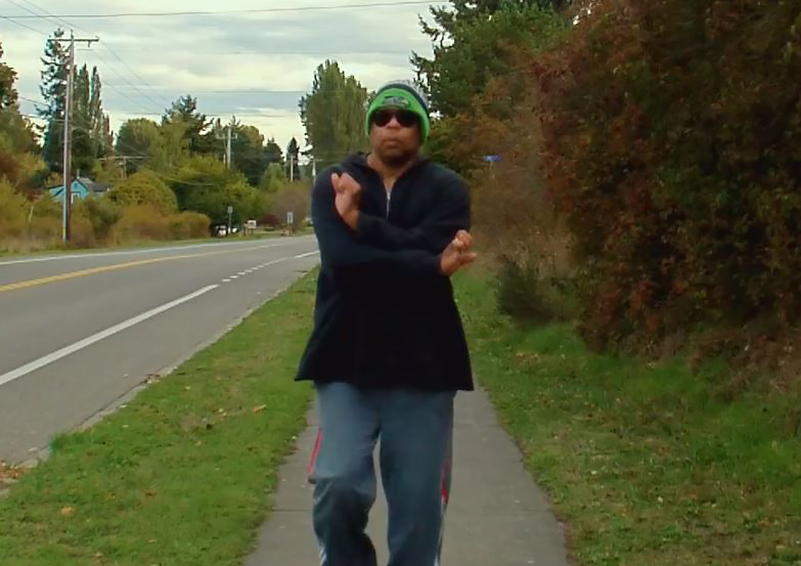 Man walking up the sidewalk in a wooded area, swinging his arms, wearing a green hat, sunglasses, dark shirt and jeans