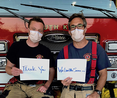 Two firemen wearing face masks sitting on the front of a red firetruck, holding signs that say "Thank You" and "We Got This Seattle"