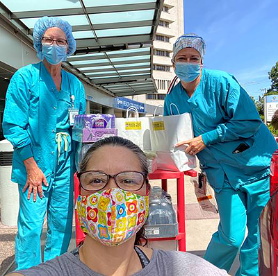Two healthcare workers in aqua scrubs and one woman in glasses and a colorful mask take a selfie in front of bags of donated food on a red rolling cart