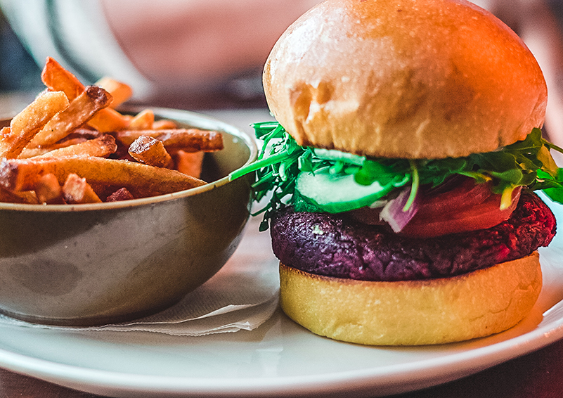 A black bean burger with tomato and spinach on a bun and a bowl of fries