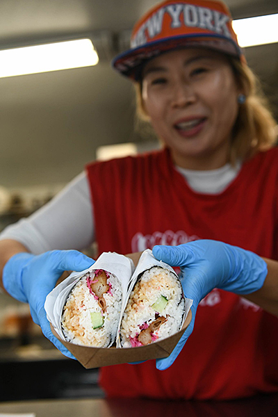 An Asian woman in a red shirt and orange NEW YORK hat holds out a to-go plate of sushi burritos