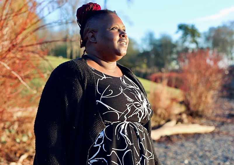 Video still of a Black woman gazing out toward a body of water on a shoreline with hope in her eyes