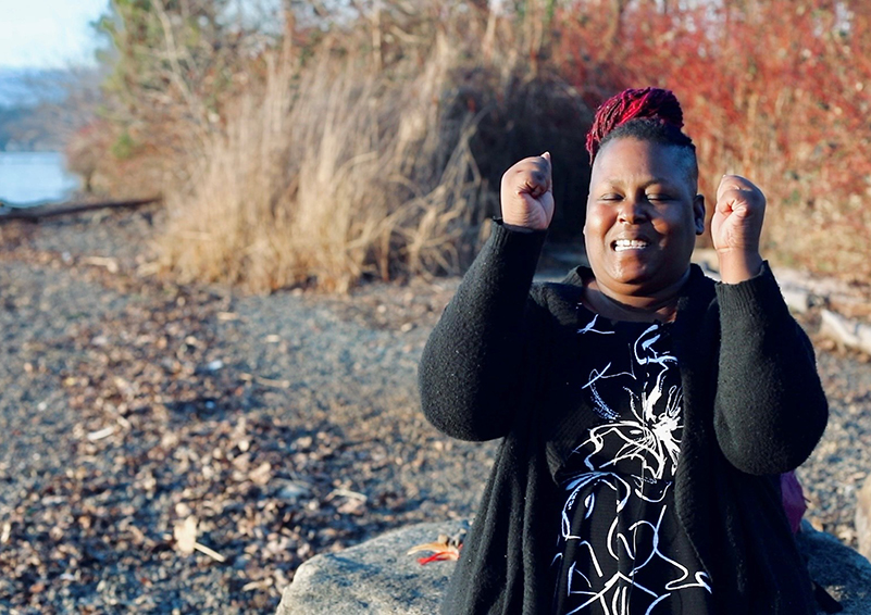 Video still of a Black woman sitting on a rock on a shoreline "dancing" as she sits