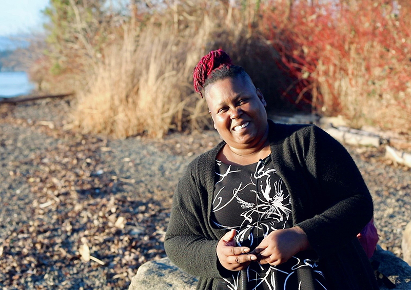 Video still of a Black woman sitting on a rock on a shoreline, smiling broadly