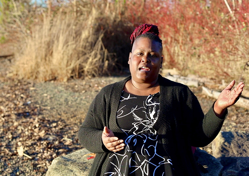 Video still of a Black woman sitting on a rock on a shoreline, gesturing with her hands as she speaks